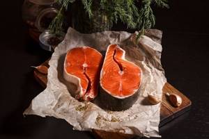 two raw chum salmon steaks on parchment paper on a cutting board, spices and garlic cloves nearby